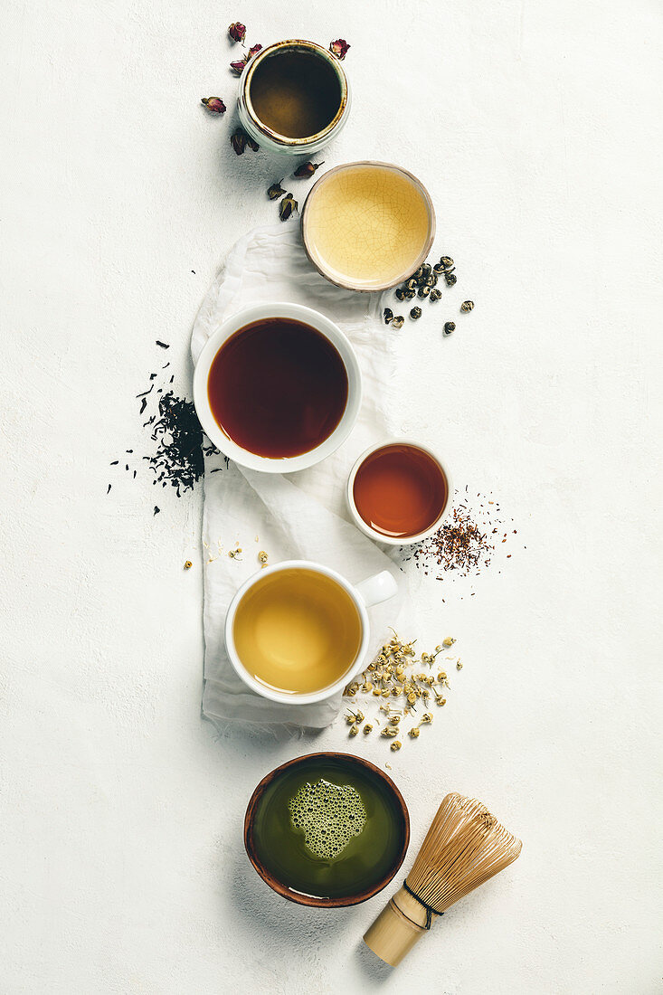 Cups of tea with aromatic dry tea in bowls on a white vintage background. Rose tea, green tea, black tea, rooibos tea, chamomile tea and matcha tea.