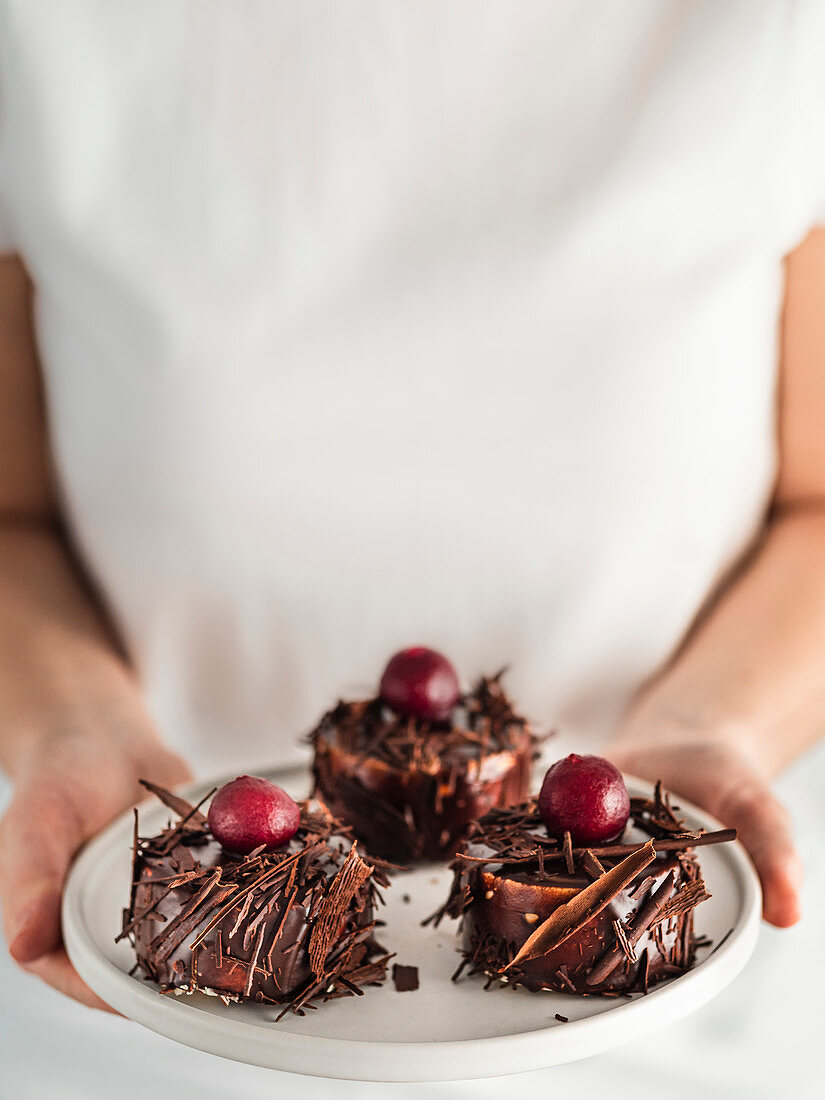 Woman in white t-shirt holds plate with three mini black forest cakes with cherry and chocolate shards. Vertical. Copy space top
