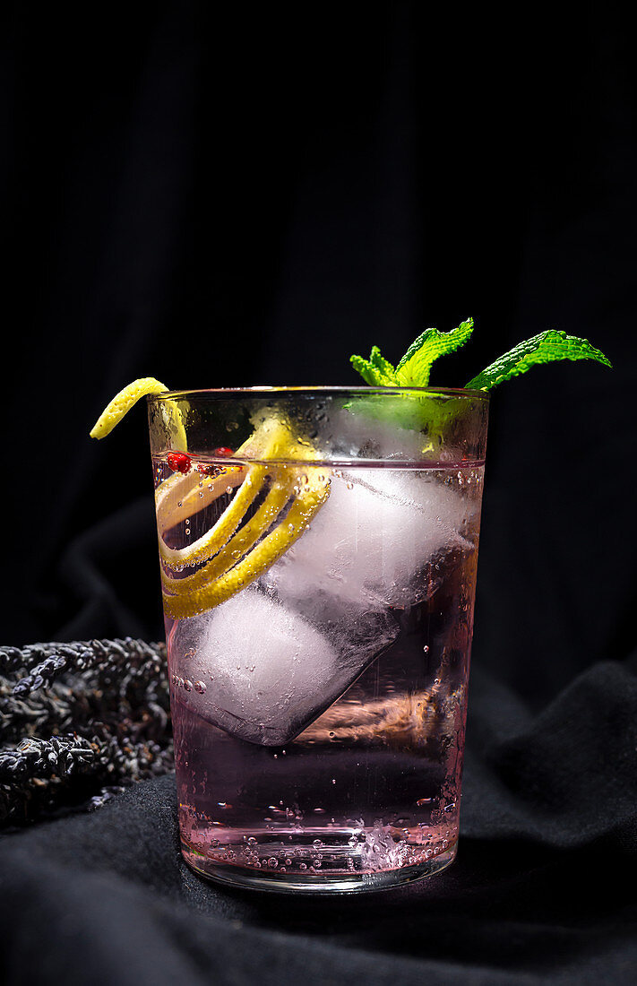 Gin tonic cocktail with pink tonic water, pink pepper, rosemary, mint, cinnamon, lemon and orange on dark background