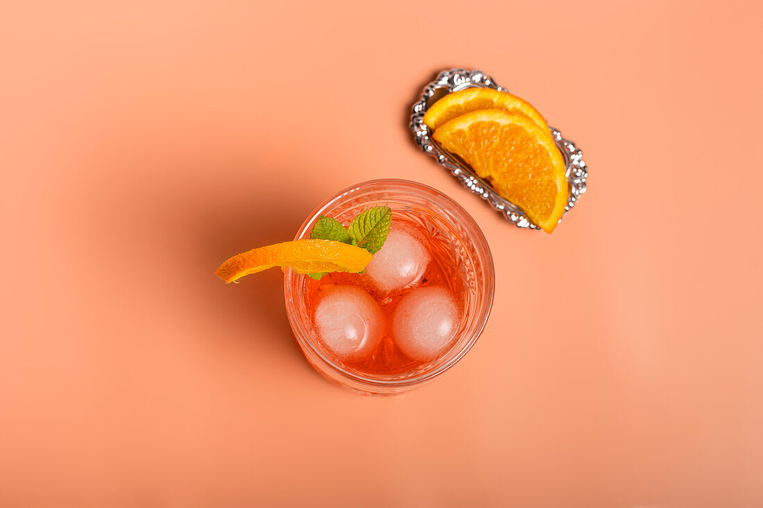 Top view of alcohol cocktail with ice cubes and sprig of mint in glass placed on pink background with orange slices