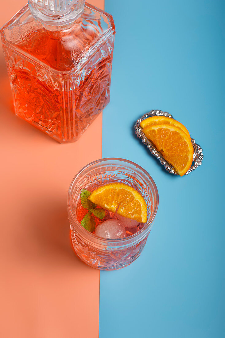 From above of glass of fresh cold cocktail with ice cubes and mint placed on colorful background with slices of ripe orange