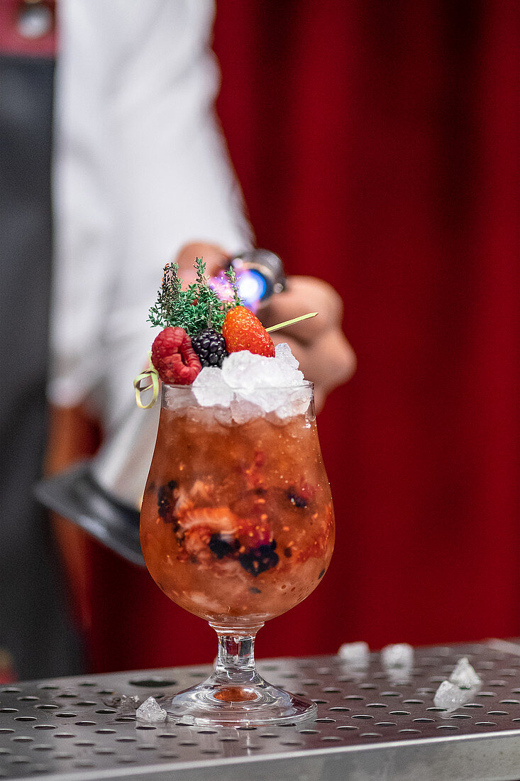 Crop anonymous bartender using burner while finishing berry cocktail with ice and fresh herbs at bar counter