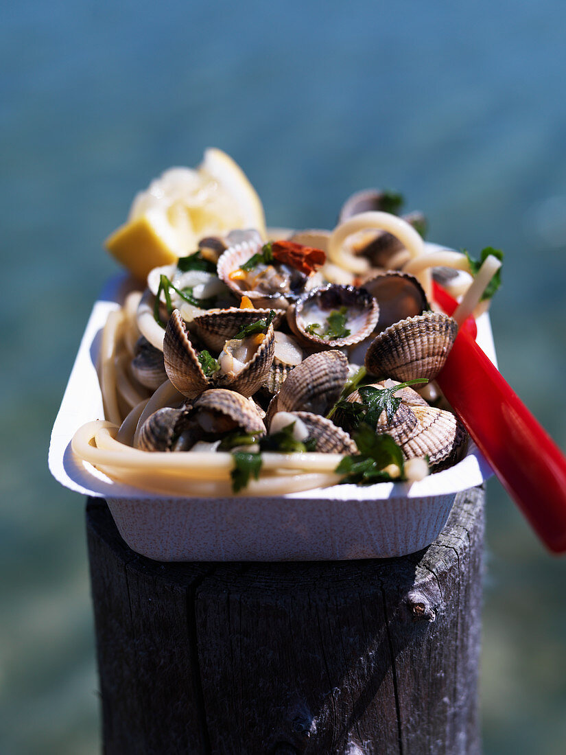 Spaghetti vongole in a cardboard bowl on a wooden post by the sea