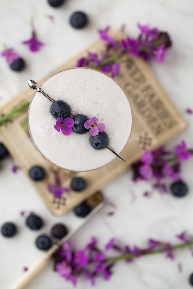 Overhead view of a cocktail with egg white foam that is decorated with blueberries and flowers