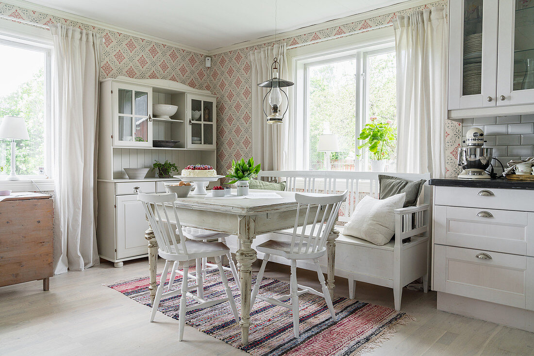 Antique, white-painted furniture, patterned wallpaper and rag rug in dining area