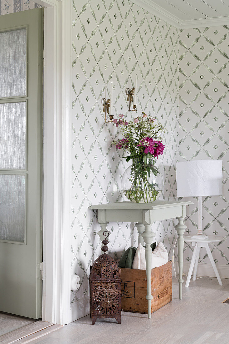 Flowers on console table and table lamp on stool against patterned wallpaper