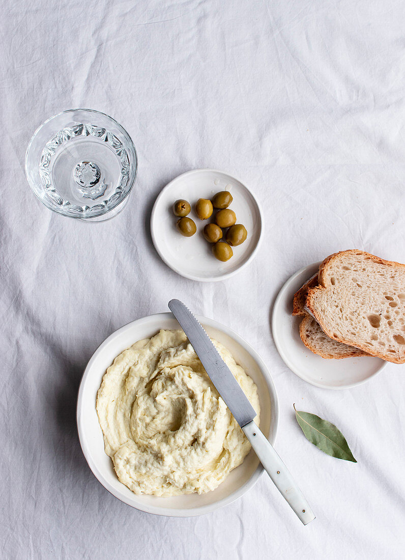 Bowl with codfish brandade, olives, bread slices and bay leaves on a kitchen table white background