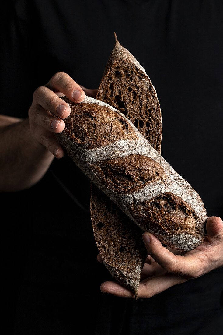 Crop male baker in apron holding cut in half loaf of fresh healthy artisan bread