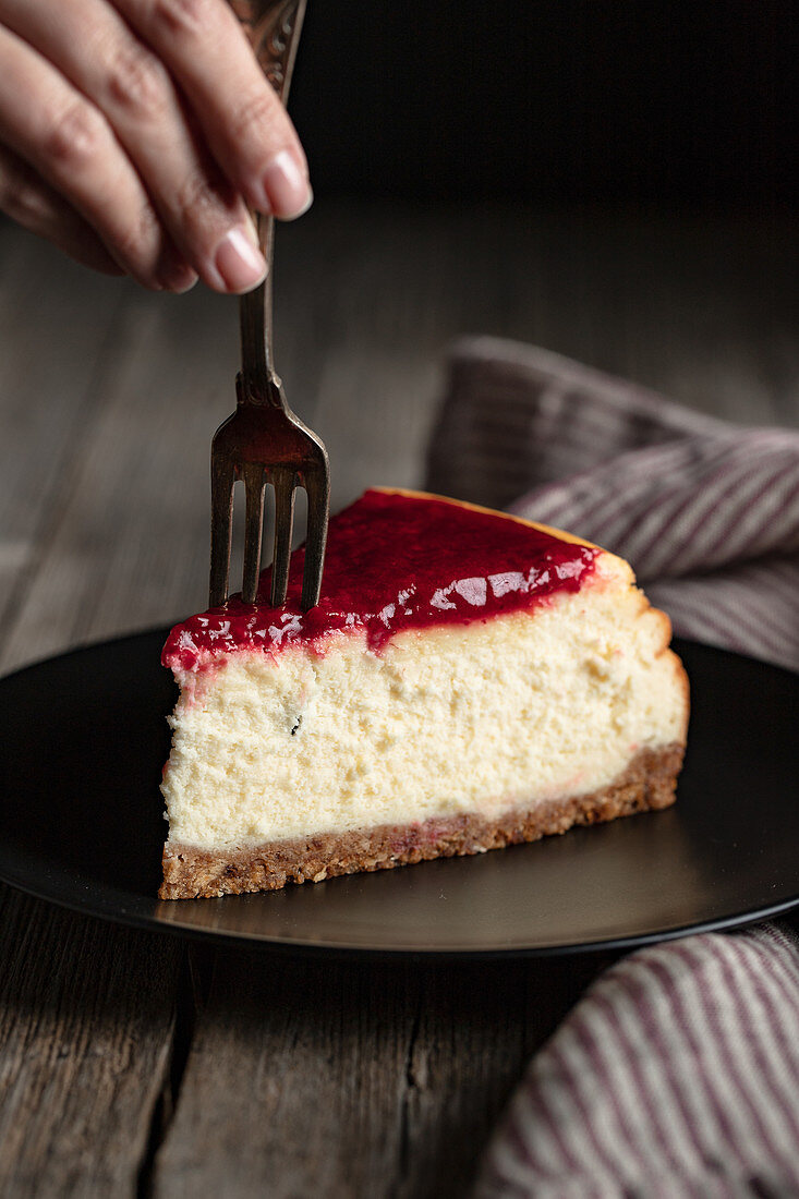 Crop female with fork eating delicious homemade cheesecake with red berry jam served on black plate on wooden table