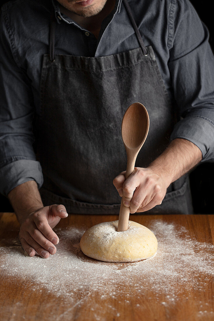 Using big wooden spoon for making hole in dough while forming artisan round bread loaf at wooden table