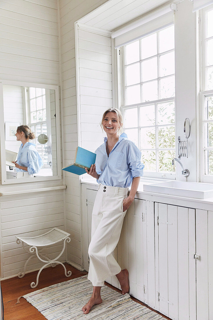 A blonde woman wearing a light-blue shirt-blouse and white trousers standing in front of a window