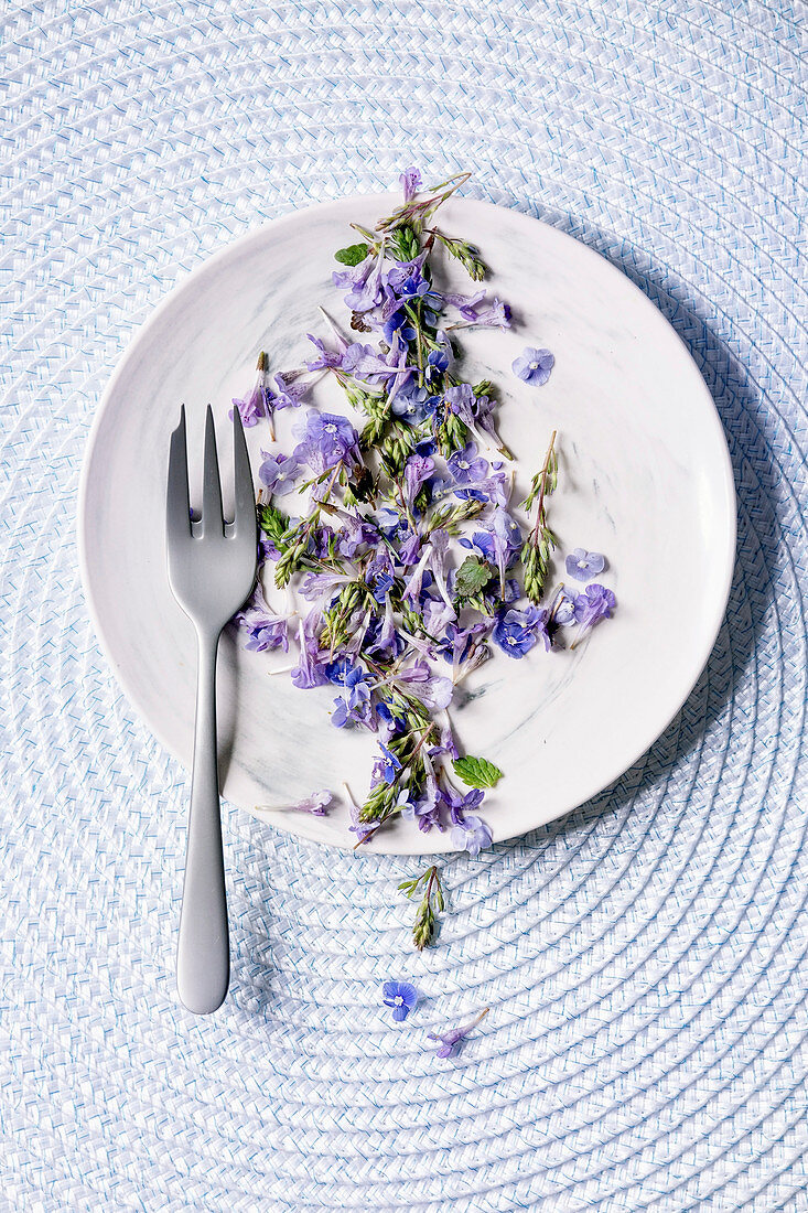 Violet purple edible flowers on white plate with dessert fork over blue knitted background