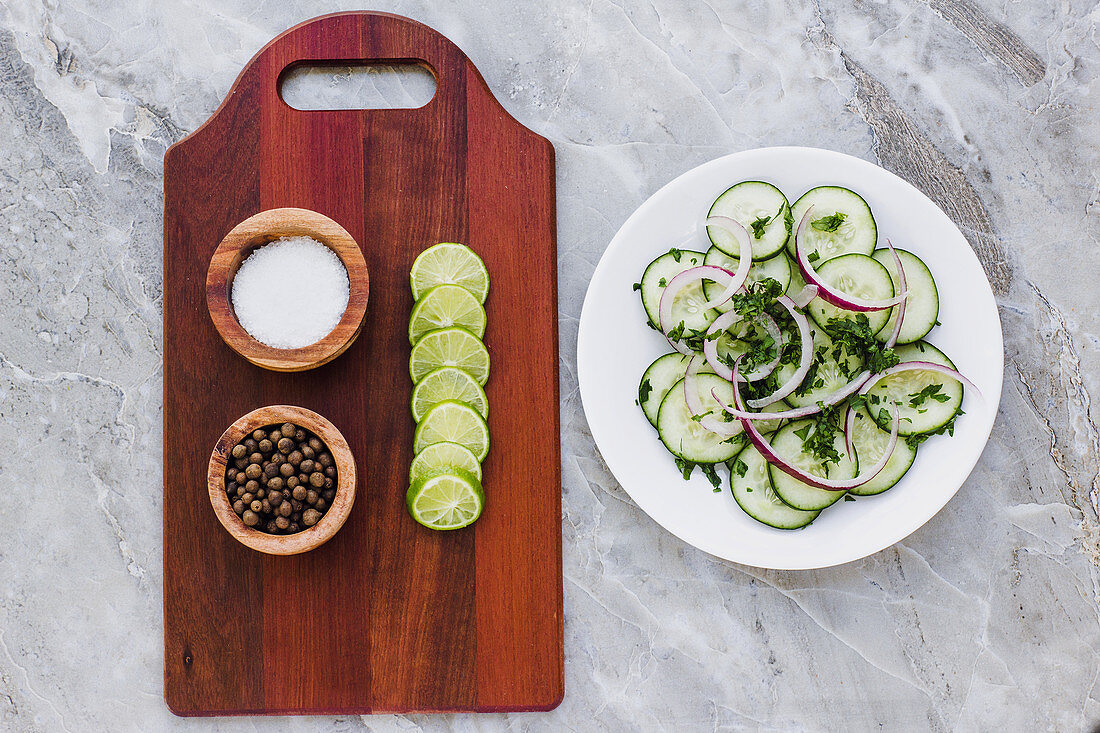 Board with salt pepper and sliced lime, plate of salad with cucumber and chopped greens with red onion