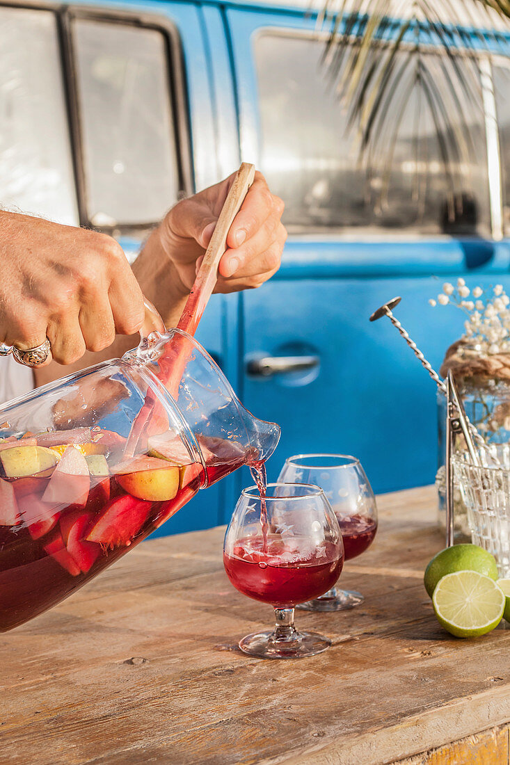 Pouring cool sangria in crystal glasses on wooden table against blue van on tropical beach