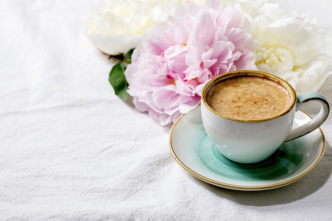 Cup of espresso coffee, pink and white peonies flowers with leaves over white cotton textile background