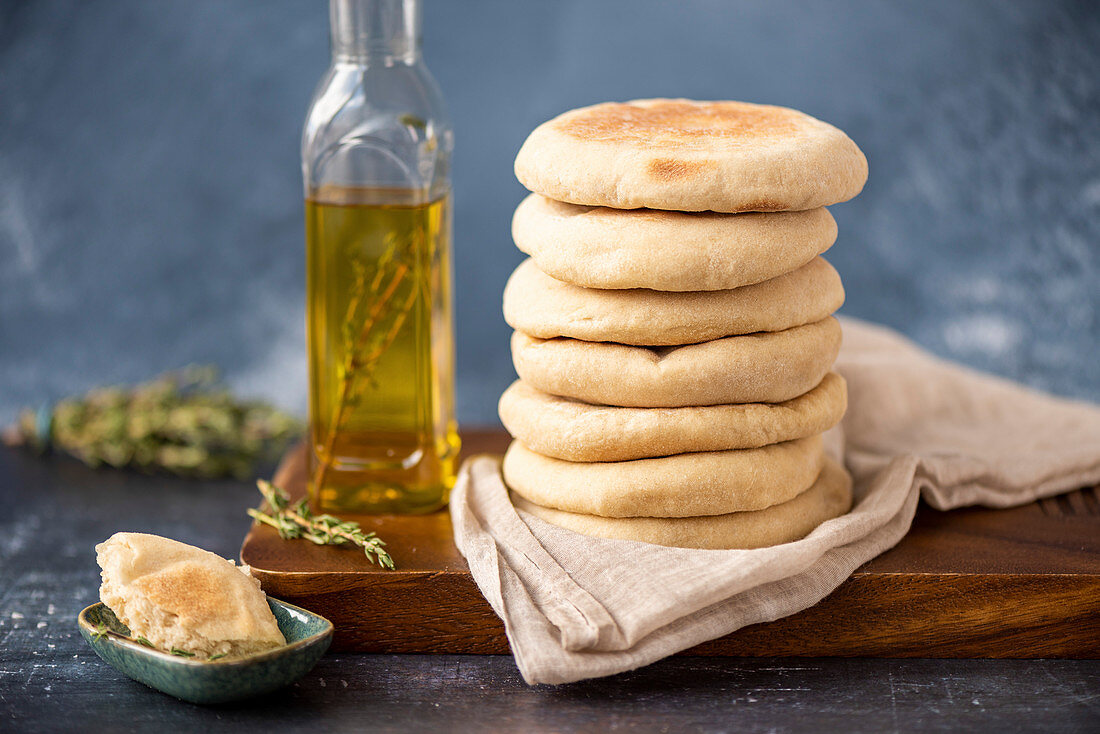 A stack of Turkish flatbread called bazlama on a wooden board, a bottle of olive oil, fresh thyme and a small bowl accompany.