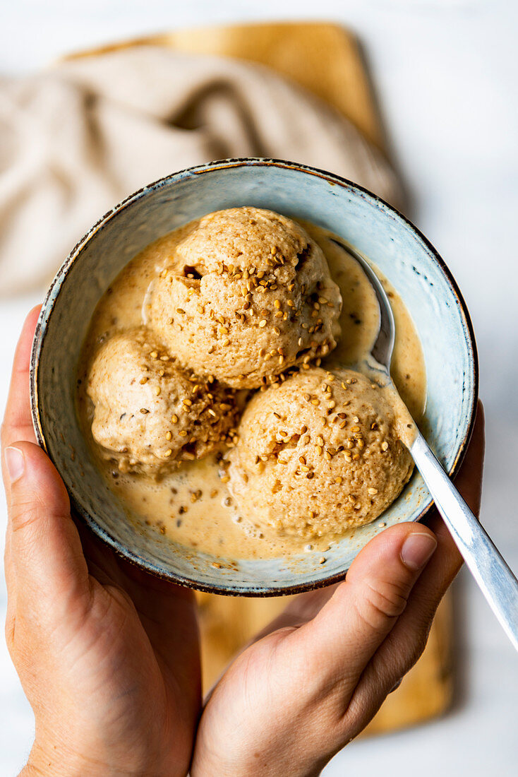 Hands holding a bowl with dollops of tahini almond milk ice cream topped with sesame seeds, a spoon in the bowl accompanies.