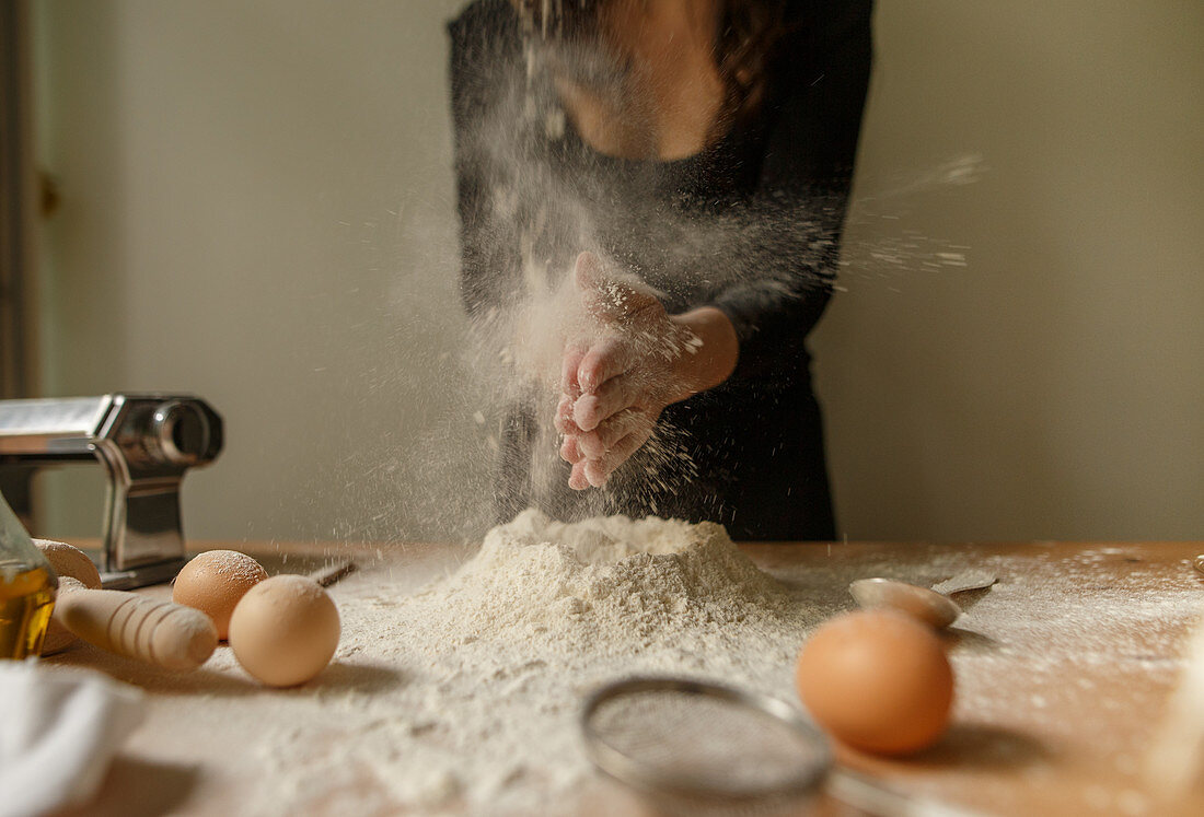 Unrecognizable female in black outfit clapping hands with flour while preparing dough on table with kitchen devices and eggs at home