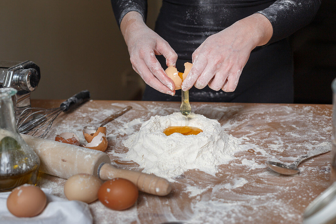 Faceless female in black leather dress breaking raw eggs into flour mass during preparation of pasta dough in kitchen at home in daytime