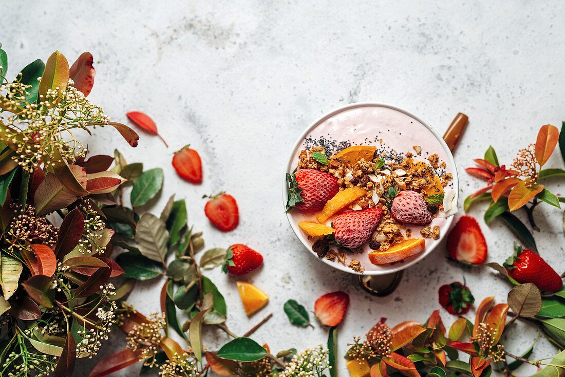 Smoothie Bowl mit Erdbeeren und Nüssen, umgeben von Blüten, Blättern und Früchten