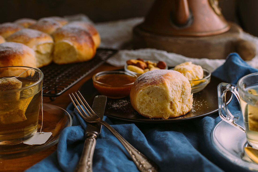 Ceramic plate with fresh bun and small bowls with mix of nuts near jam and butter close to cup of green tea on crumpled towel