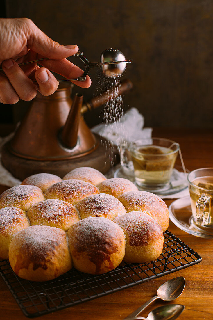 Crop anonymous chef sifting icing sugar using small metal sieve while decorating tasty golden buns near cups with green tea bags on table