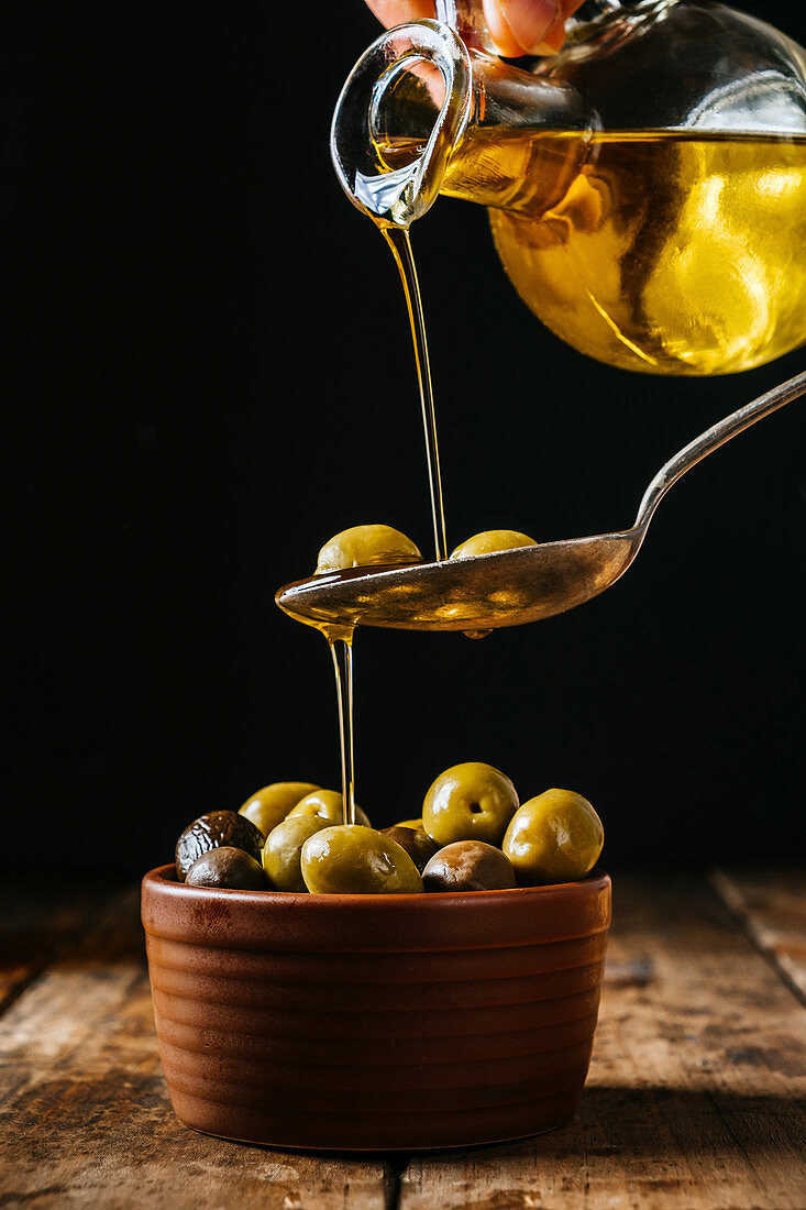 Pouring oil in spoon and ceramic bowl with olives placed on wooden table in kitchen