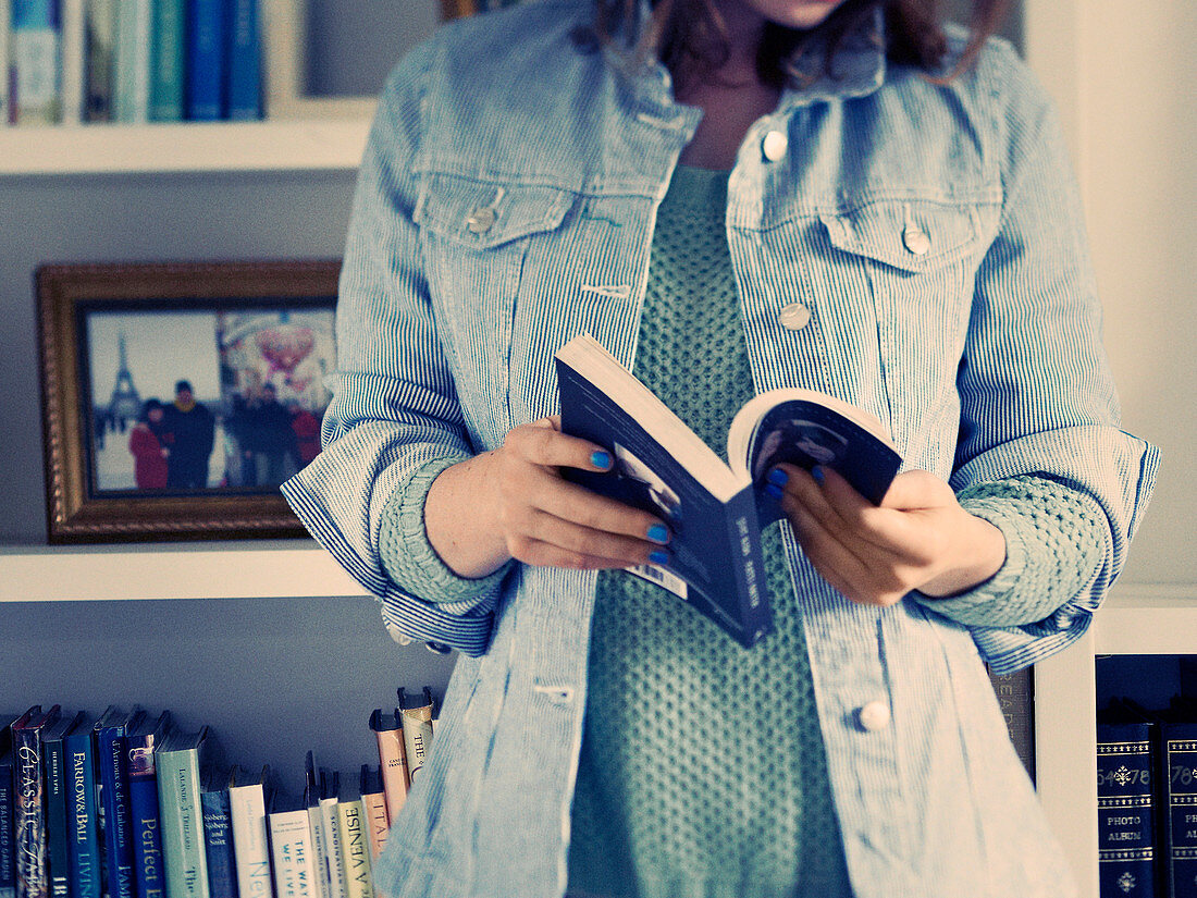 A woman wearing a denim shirt and holding a book standing in front of a bookshelf