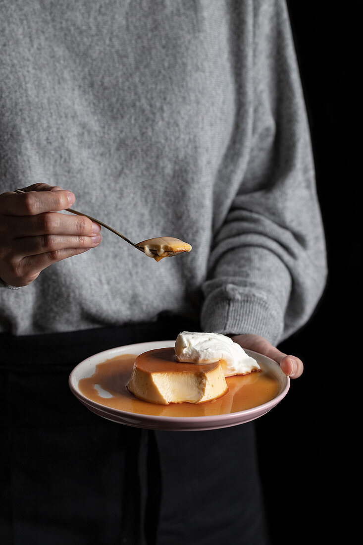 Unrecognizable female eating delicious caramel custard placed on plate with whipped cream on black background