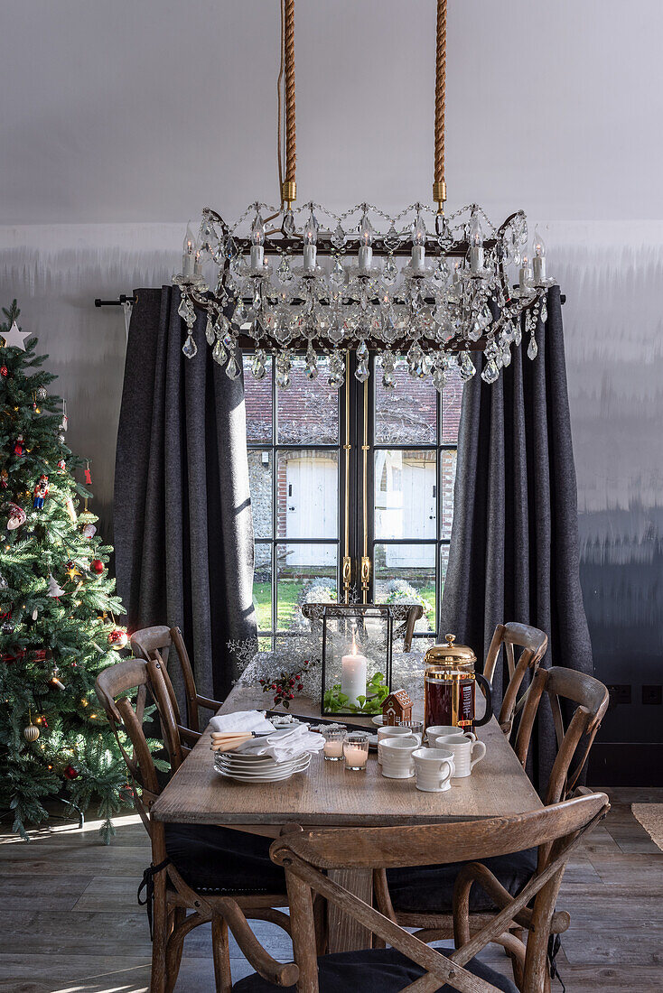 Rustic wooden table set with tea service and lantern below chandelier with Christmas tree in corner of room