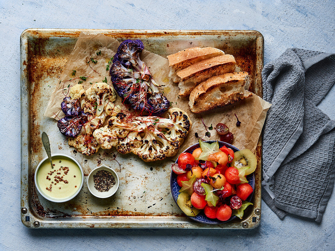 Roasted cauliflower on a baking tray with a melon salad, bread and a creamy dip