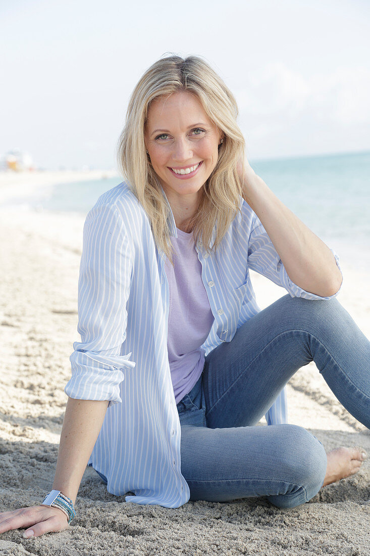A blonde woman by the sea wearing a blue-and-white striped shirt and jeans