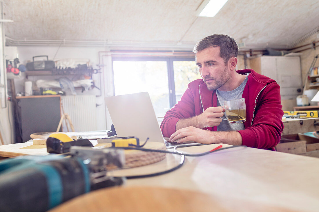 Male carpenter drinking tea and working at laptop