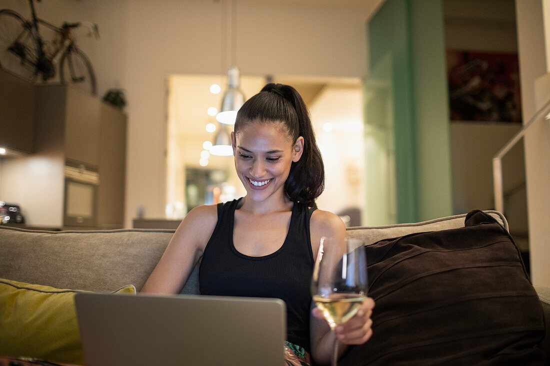 Smiling woman using laptop and drinking white wine