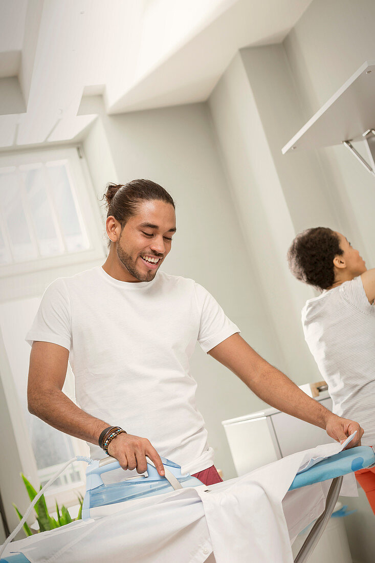 Young man doing laundry, ironing shirt in laundry room