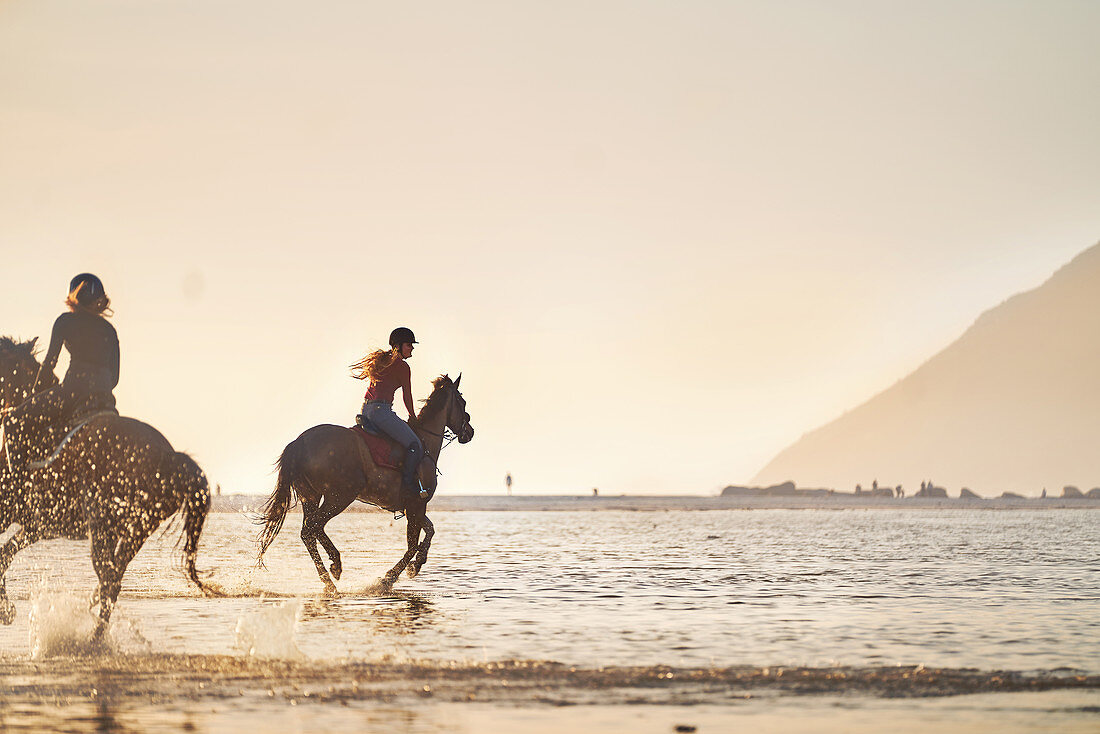 Young women horseback riding in ocean surf at sunset