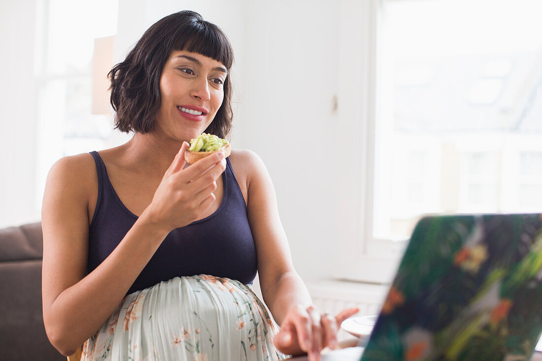 Happy pregnant woman eating avocado toast at laptop