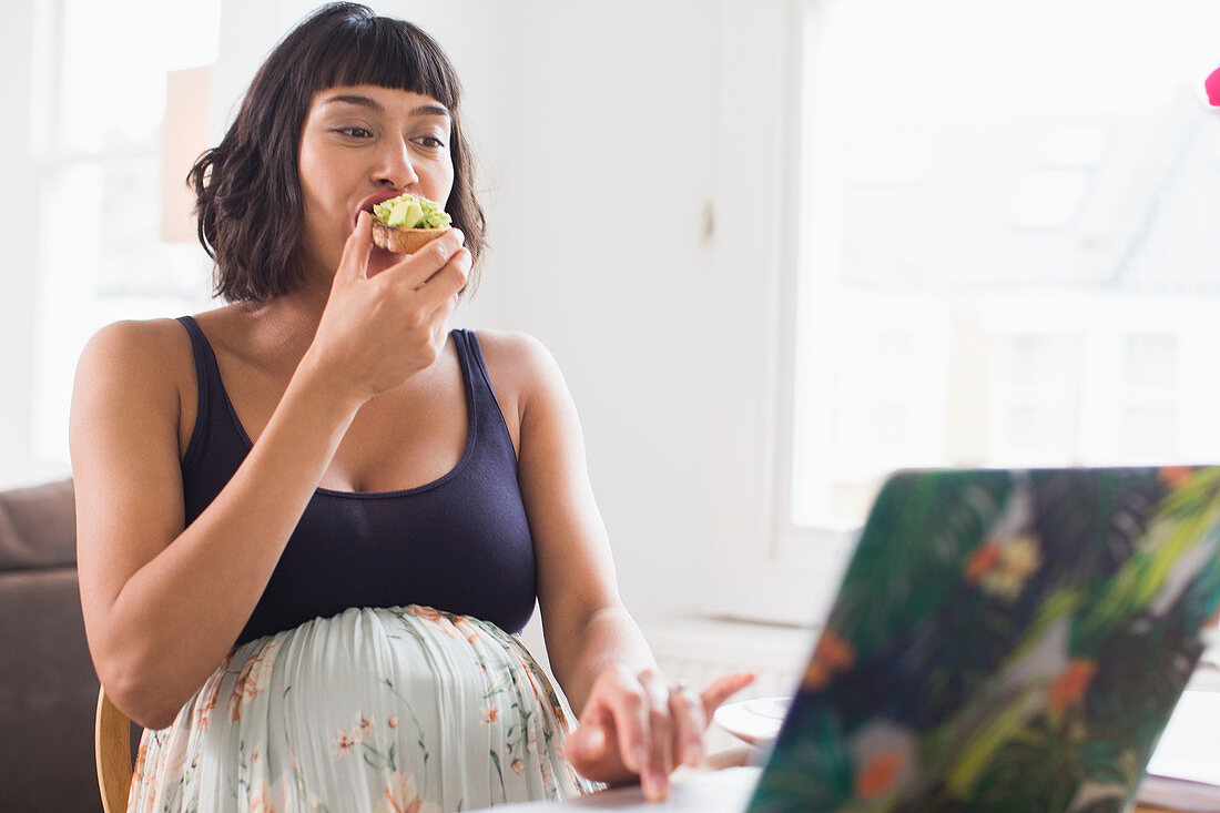 Pregnant woman eating avocado toast at laptop