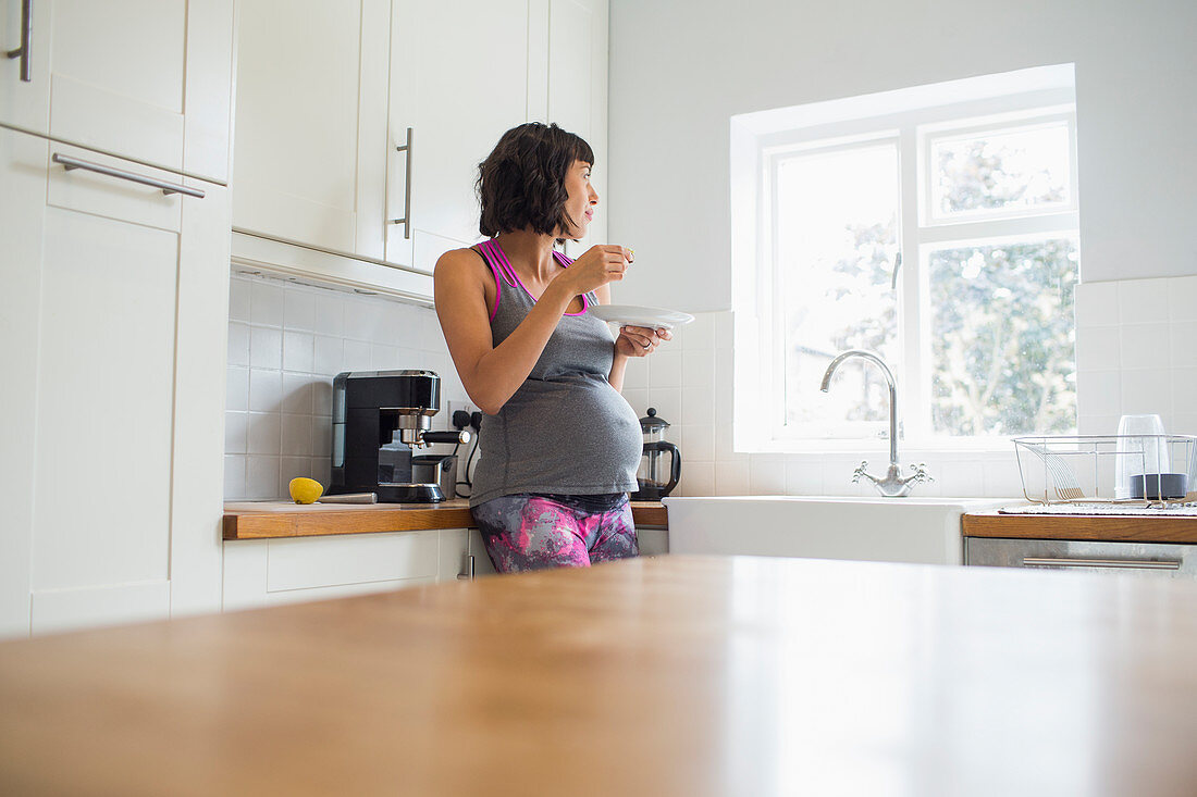 Pregnant woman eating in kitchen looking out window