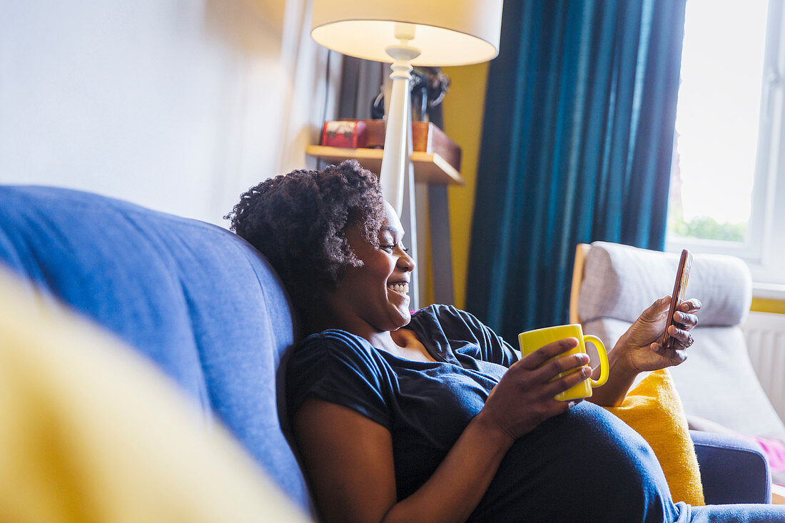 Pregnant woman relaxing on sofa with tea and smart phone