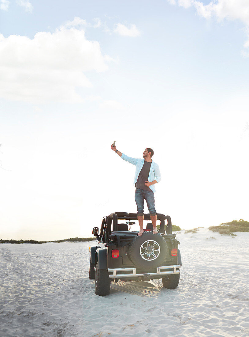 Young man phone taking selfie on top of jeep tire on beach