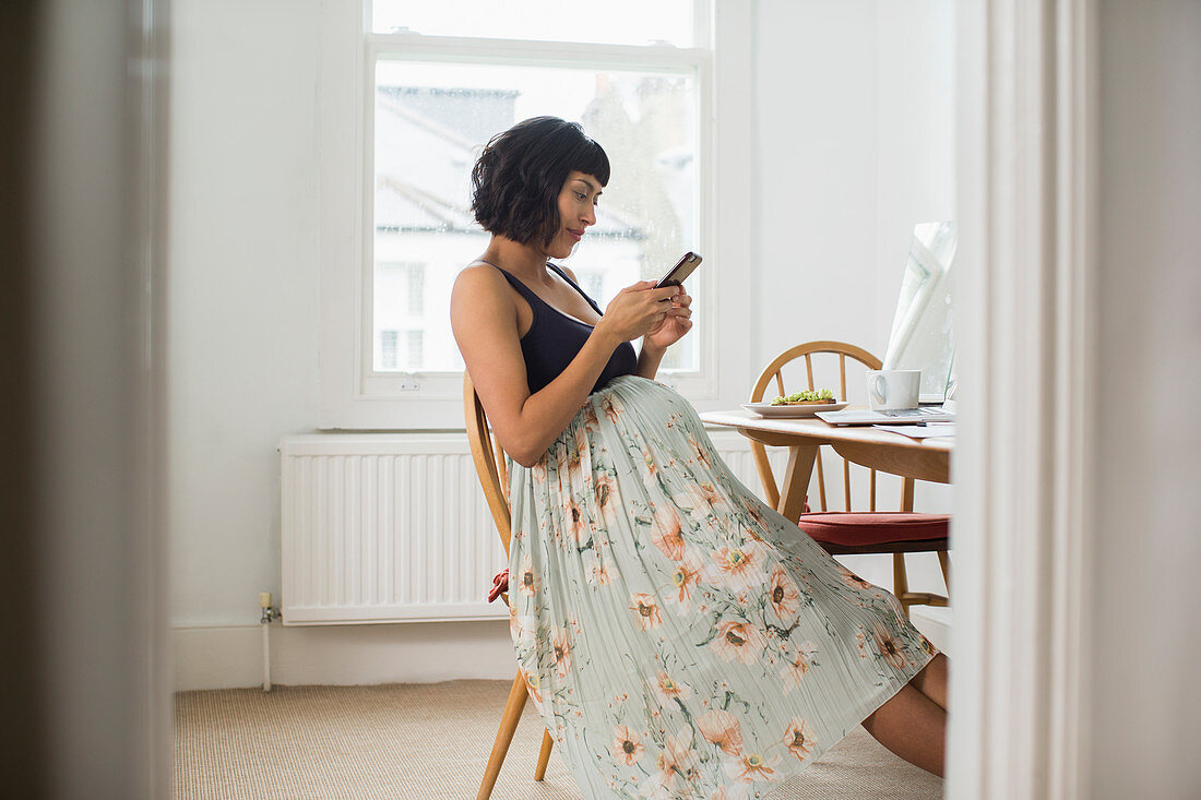 Pregnant woman in floral dress using smart phone at table