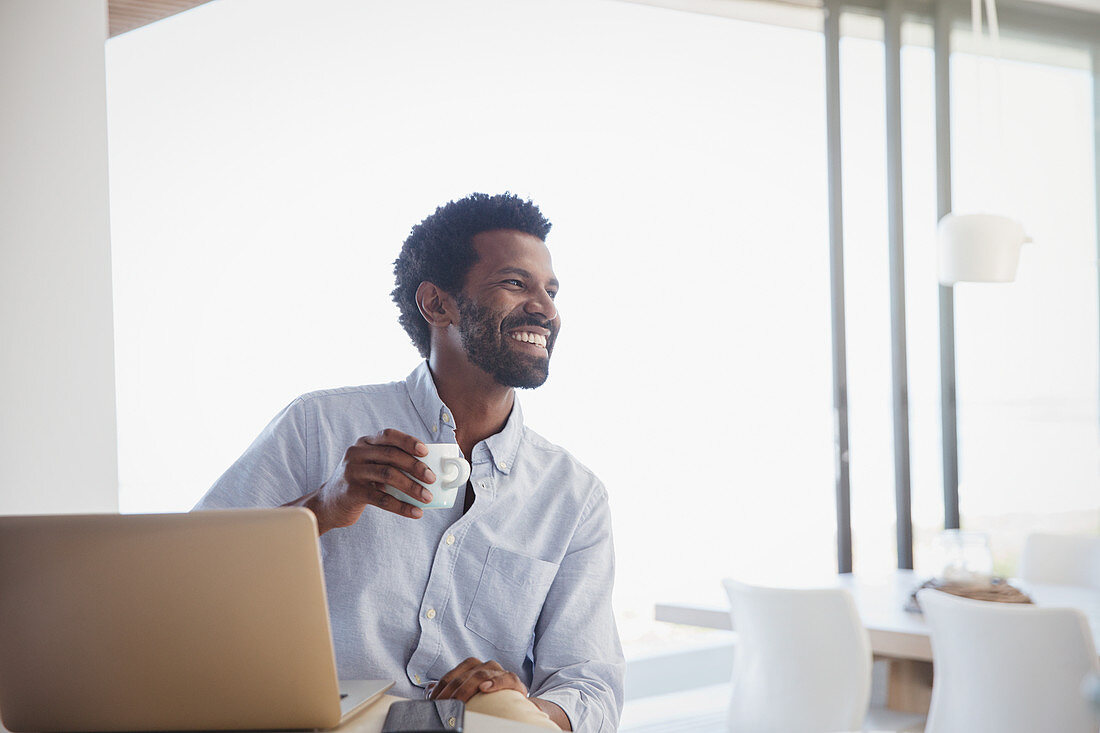 Smiling man drinking coffee and working at laptop at home