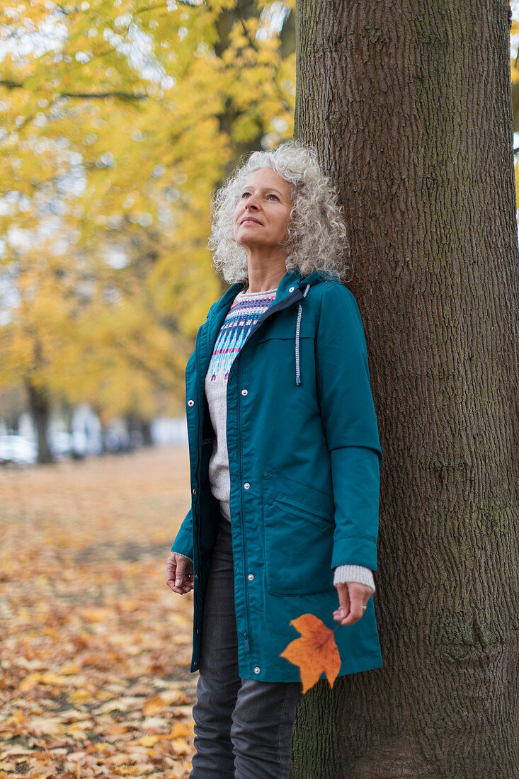 Serene senior woman leaning against tree in autumn park