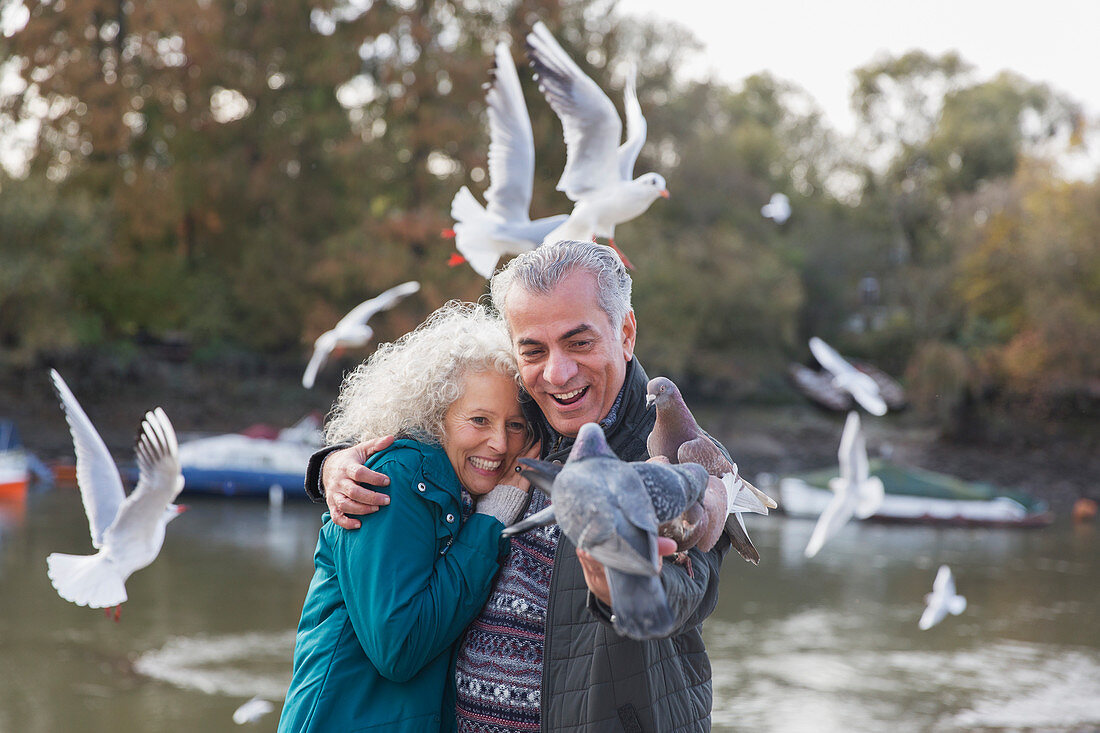 Playful senior couple feeding pigeons at pond in park