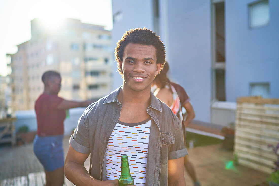 Confident young man drinking beer on sunny urban rooftop