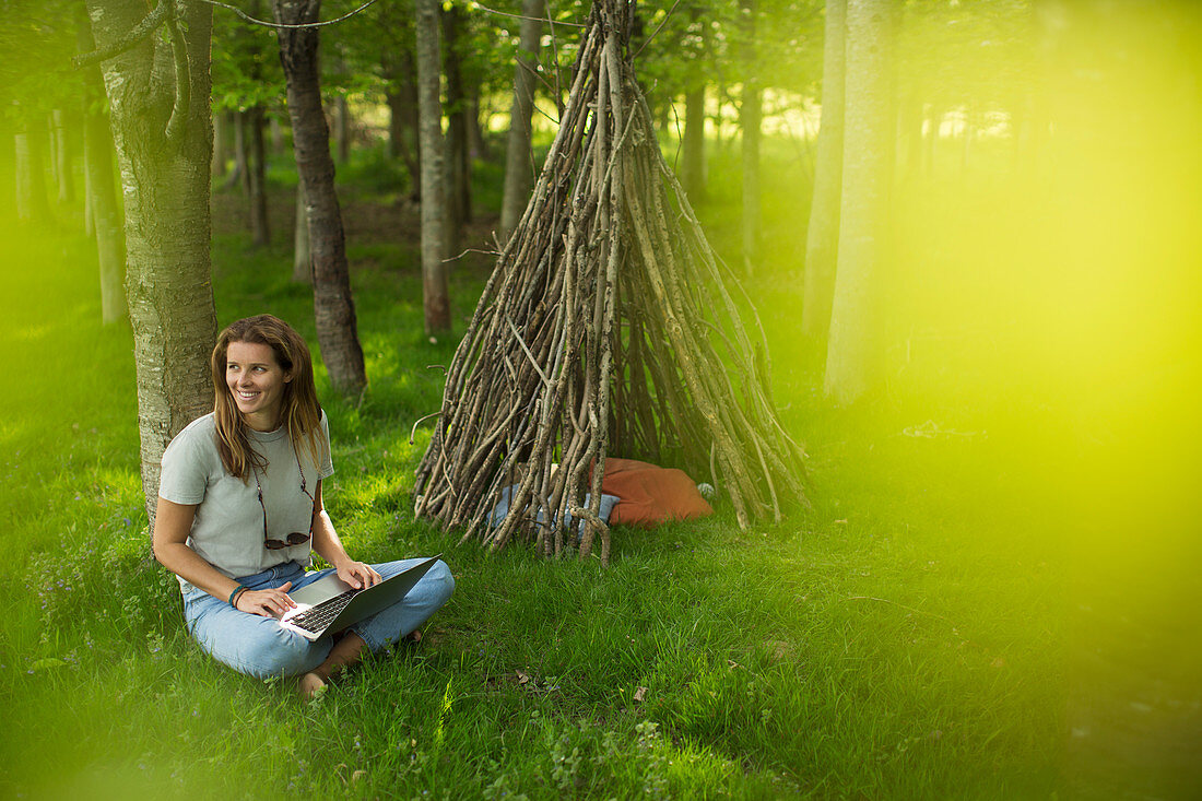 Smiling woman using laptop outside branch teepee in woods