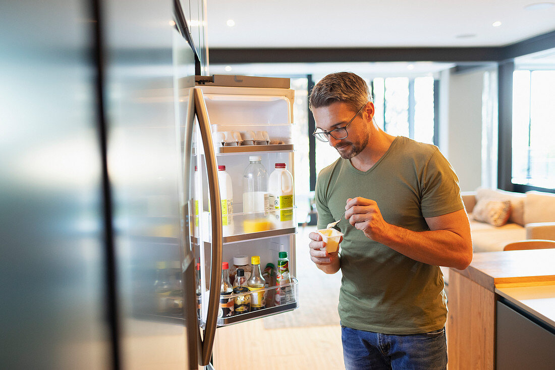 Man eating yogurt at open refrigerator in kitchen