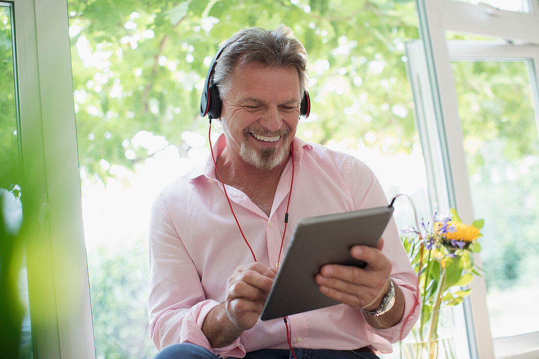 Senior man using headphones and tablet at window