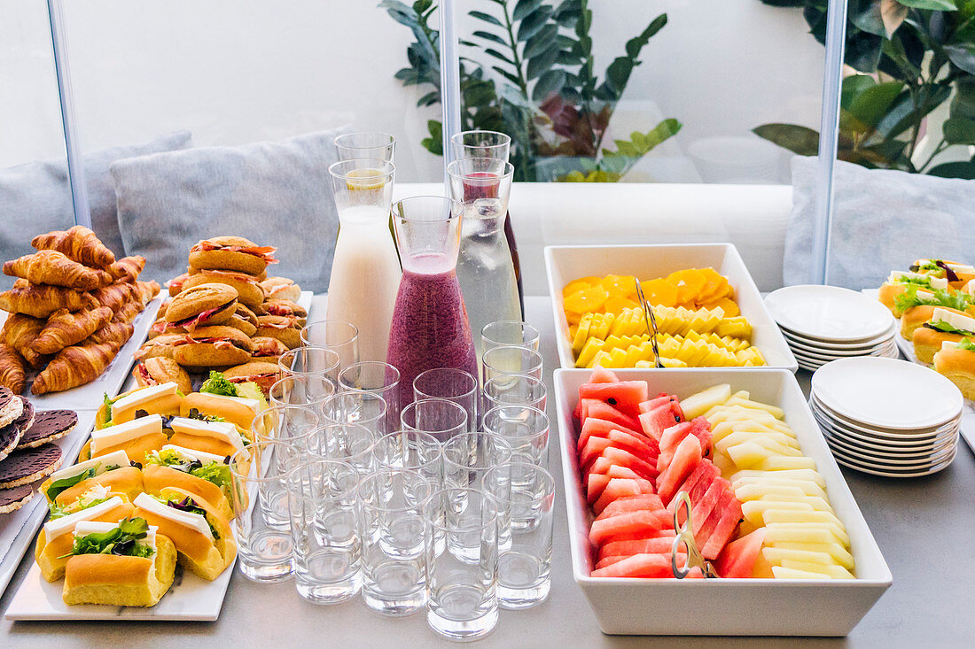 Assorted smoothie in glass bottles and sandwiches with slices of bacon and sweet croissants, empty glasses for party